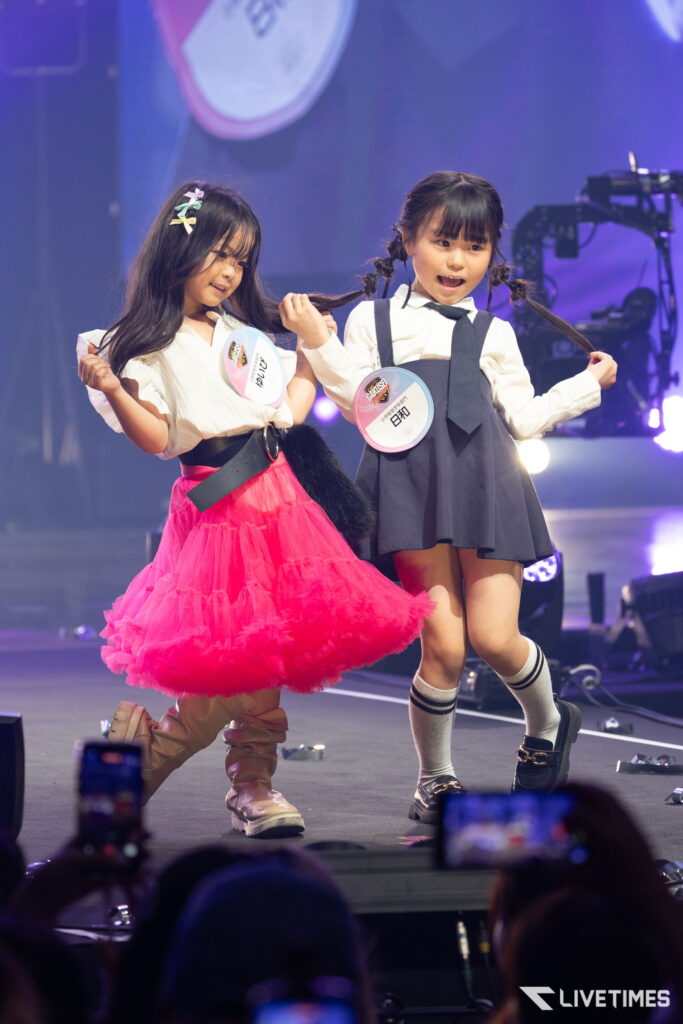 Two young girls perform on stage, holding braided hair, one in a pink tulle skirt and the other in a grey uniform, with badges visible.
