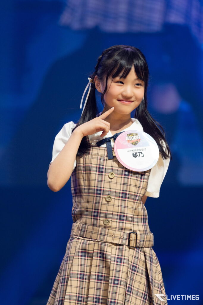 Smiling young girl on a stage wearing a beige plaid dress with a round name badge, posing with her finger to her cheek against a blue background.