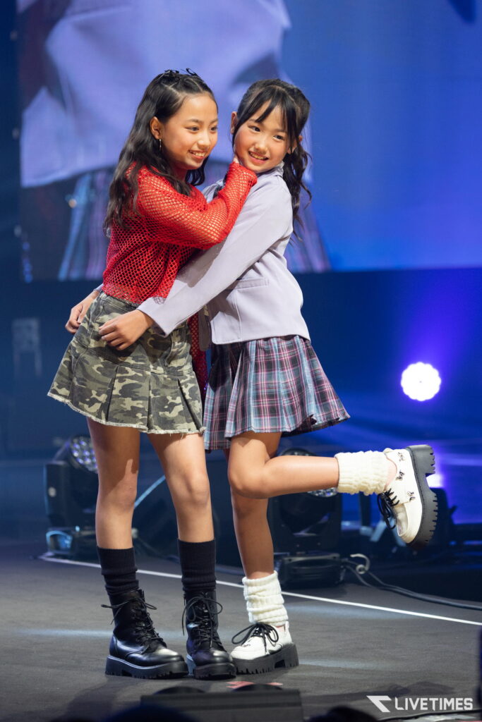 Two young girls embrace and smile on a stage, wearing plaid skirts and chunky boots with blue stage lights behind them.