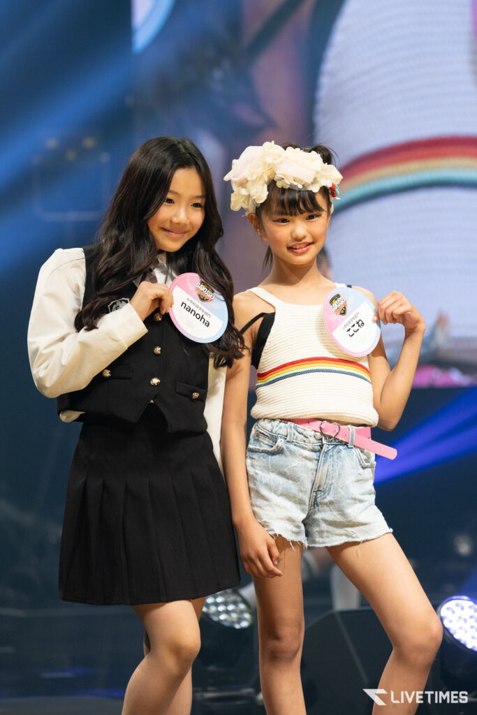 Two girls posing on stage, one in a black vest and white shirt with a badge, the other in a white knit top and denim shorts with a flower headband and badge.