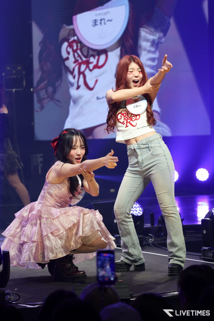 Two female performers dance on a lit stage; one stands in light blue jeans and a white cropped shirt, pointing outward, while the other kneels in a pink frilly dress with a hair bow.