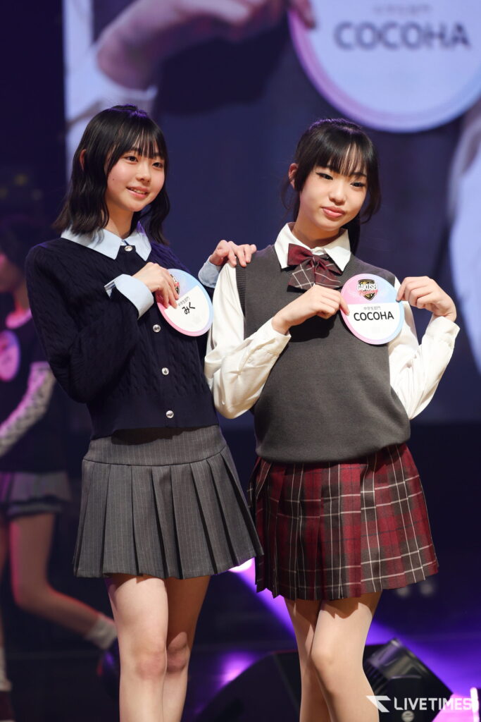 Two young girls in school uniforms pose on stage, each wearing a round name badge on their chest.