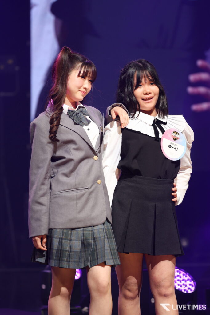 Two young girls in school uniforms posing on a stage with purple lighting, one with a braided ponytail and the other with a name badge on her chest.