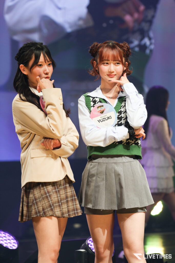 Two young women on a stage posing with hands to their chins; one in a beige blazer and plaid skirt, the other in a white shirt with a green argyle vest and gray skirt, wearing a name badge reading YUZUKI.