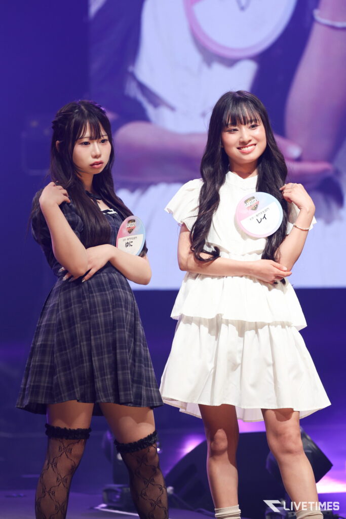 Two young women stand on a stage posing with hands on their hips; one wears a dark plaid dress, the other a white dress, both with circular event badges visible on their chests.