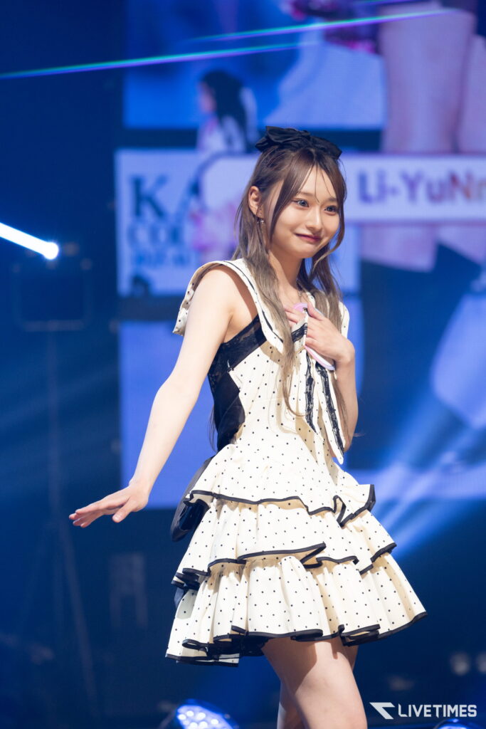 Young female performer on stage in a white polka-dot dress with black trim and a bow, smiling as she performs under blue lights.