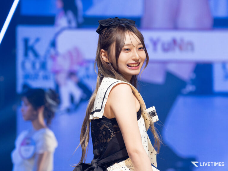 Smiling female performer on stage in a white polka-dot outfit with a black lace panel and large bow in her hair, stage lights in the background, Livetimes watermark.