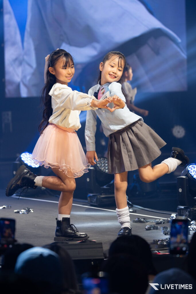Two young girls in pastel skirts dancing on a stage, holding hands and smiling, with an audience in the foreground.