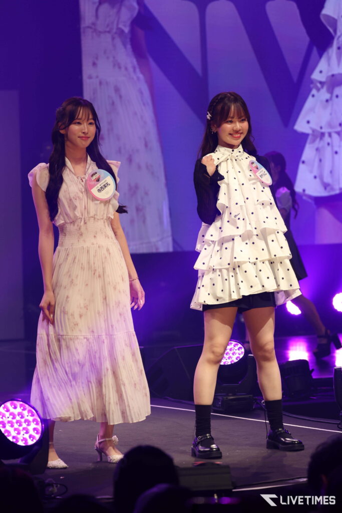 Two young women on a lit stage wearing pastel dresses with badges, smiling at the audience.