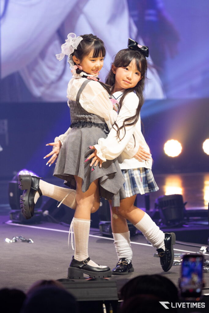 Two young girls in plaid skirts perform on stage, embracing mid-dance under bright stage lights.