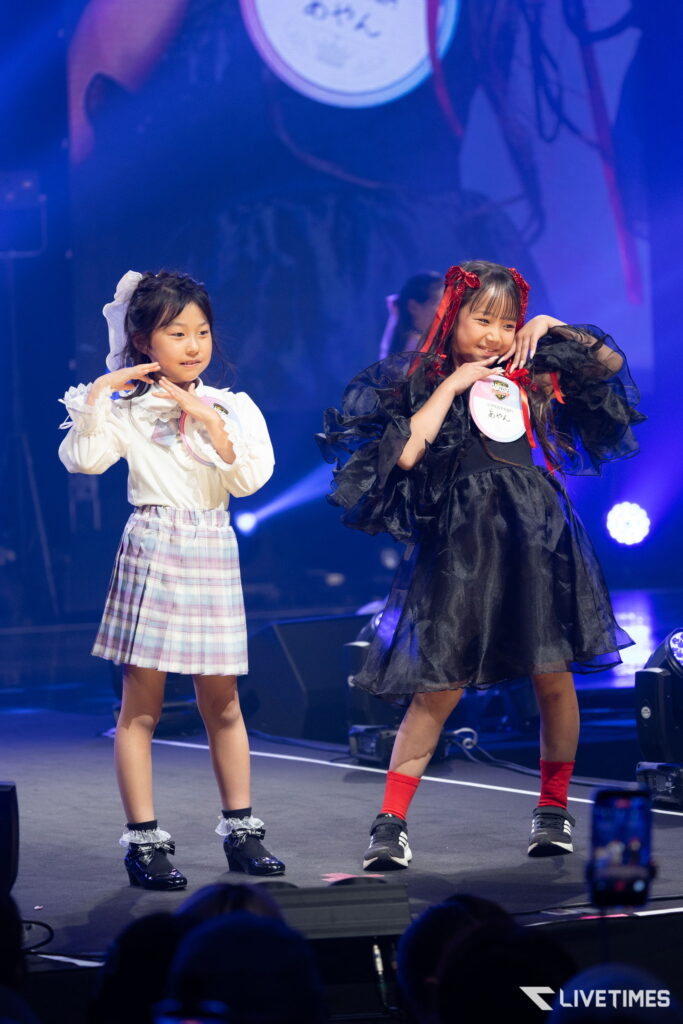 Two young girls performing on a stage: one in a white plaid dress with a bow, the other in a black frilly dress with red ribbons, posing with hands near their faces.