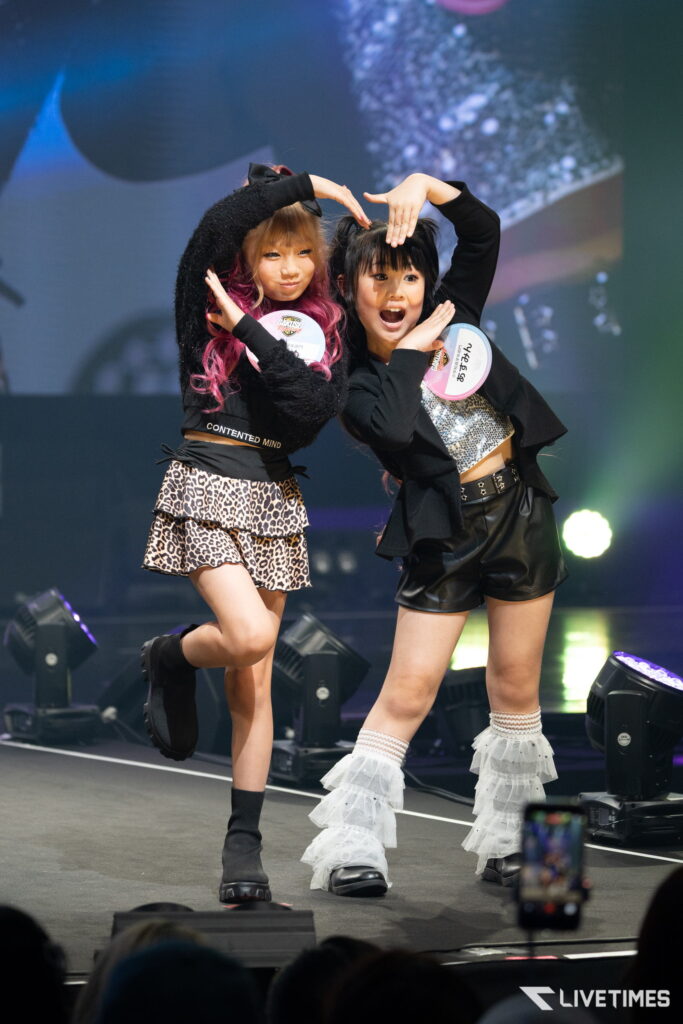 Two young dancers perform on stage, posing with arms overhead and leopard-print skirts, badges on chests.