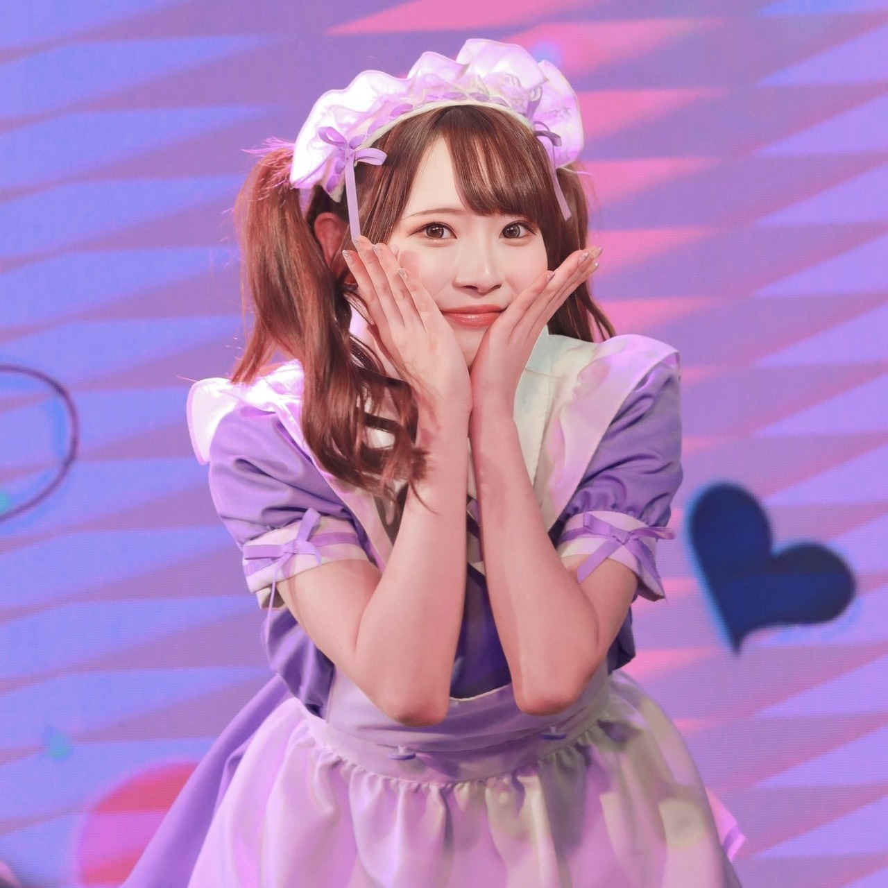Young woman in a lilac maid costume on stage, hands cupping her cheeks and smiling at the camera.