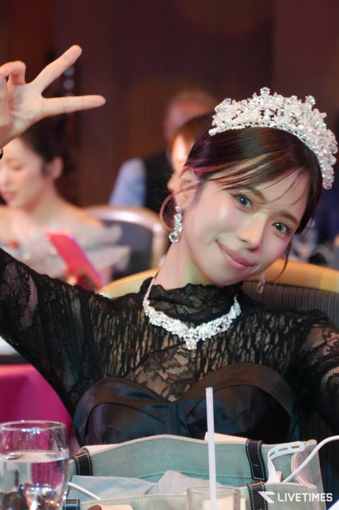 Young woman wearing a jeweled tiara and black lace dress, smiling at the camera with a peace sign gesture zone left.