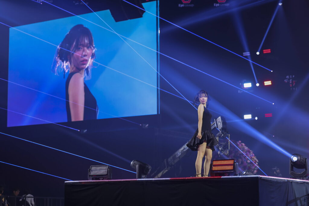 Singer in a black dress walks the catwalk during a concert, with blue stage lights and laser beams crossing the scene, and a large screen showing her portrait behind her.