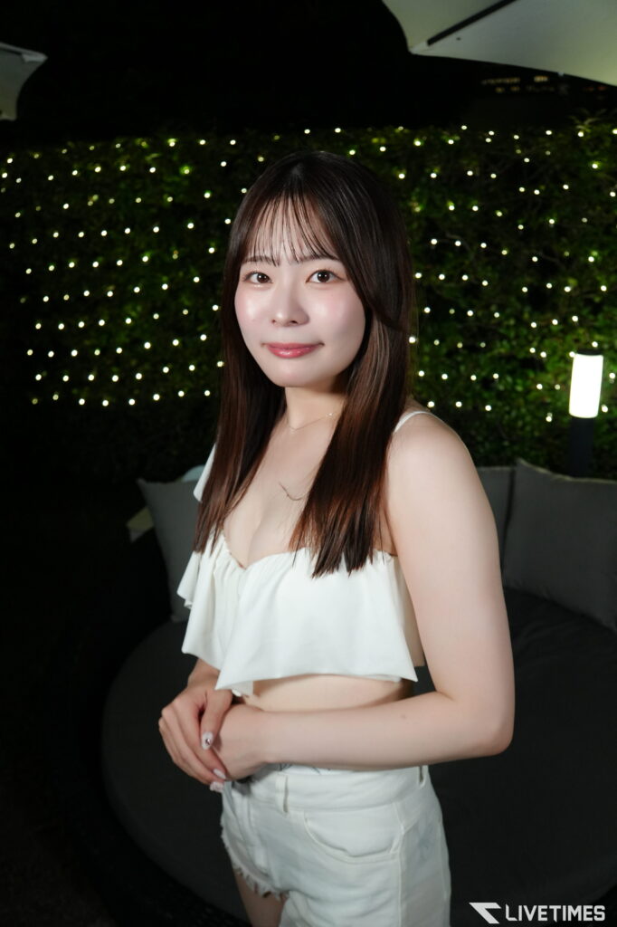 Portrait of a young woman in a white sleeveless top standing at night with a wall of twinkling lights behind her.