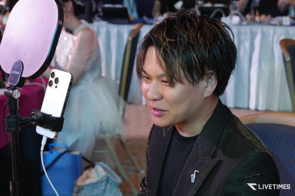 Young man with highlighted hair sits at a setup with a smartphone on a tripod and a circular ring light nearby.