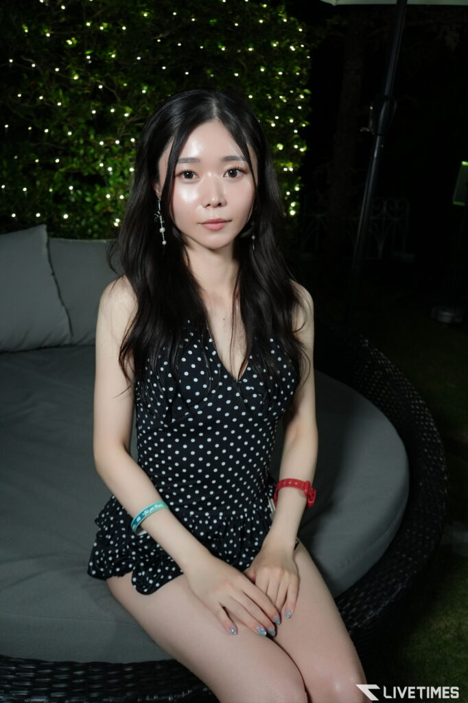 Young woman with long dark hair in a black polka-dot dress sits on a wicker couch outdoors at night, with string lights in the background.
