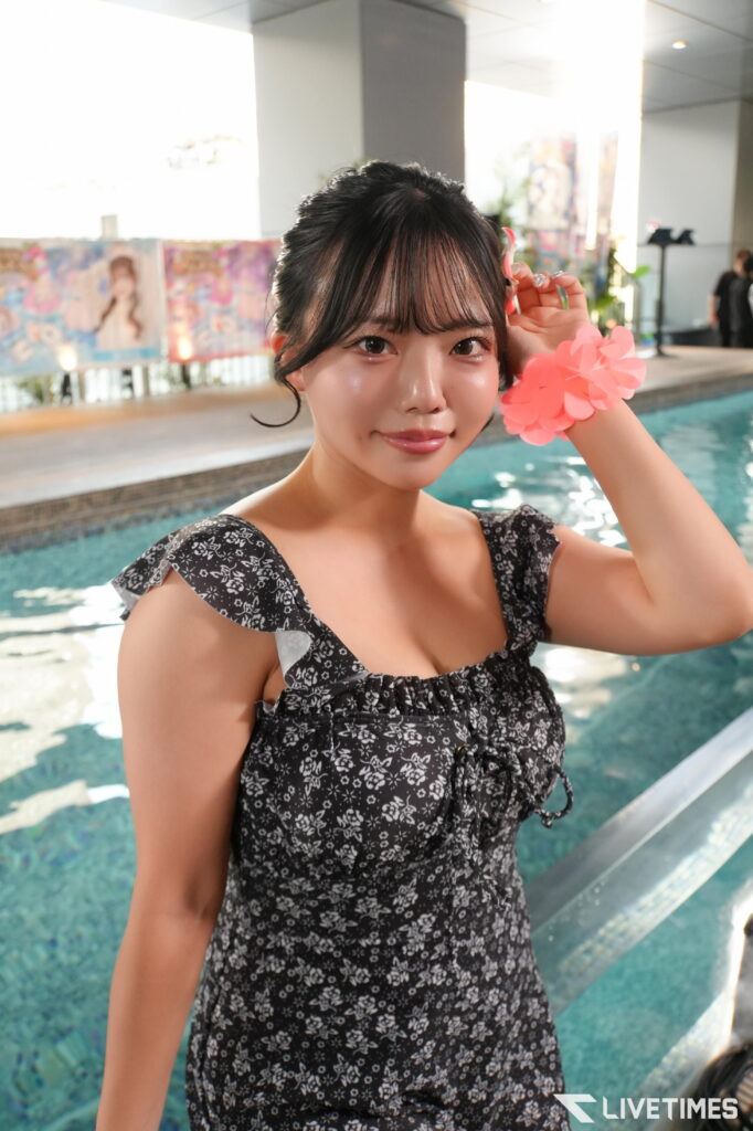 Young woman in a black floral dress by an indoor pool, adjusting a pink flower bracelet near her ear.