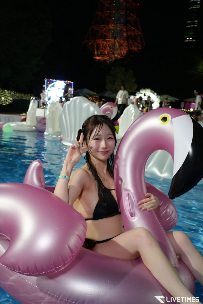Young woman in a black bikini on a pink inflatable flamingo in a nighttime pool party scene.