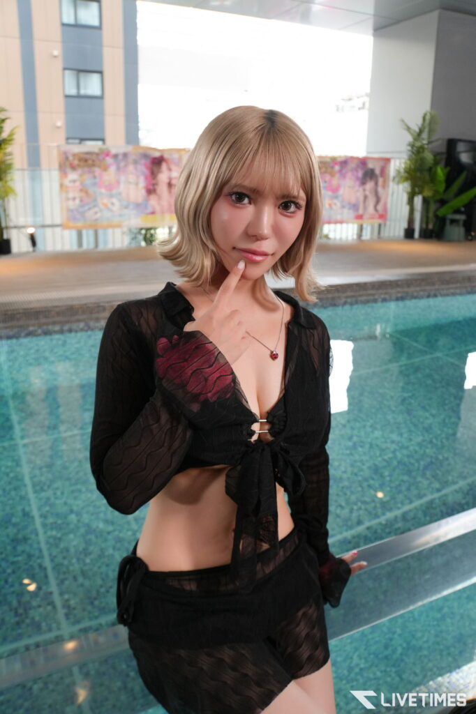 Woman with blonde hair posing by a pool, touching her chin with one finger, wearing a sheer black outfit and a red pendant necklace.