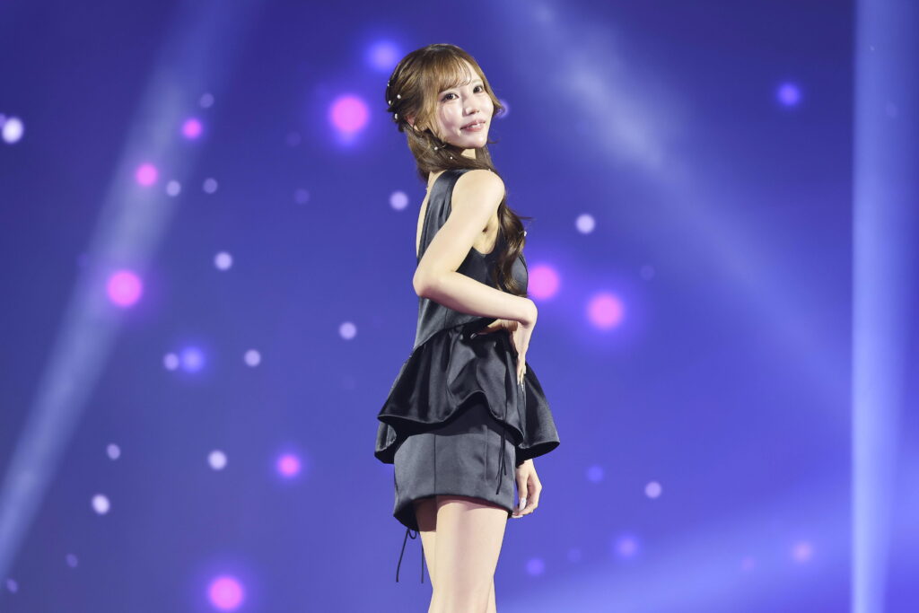 Female performer in a black satin dress posing on stage, looking over her shoulder with a smile, against a blue-purple light-filled background.