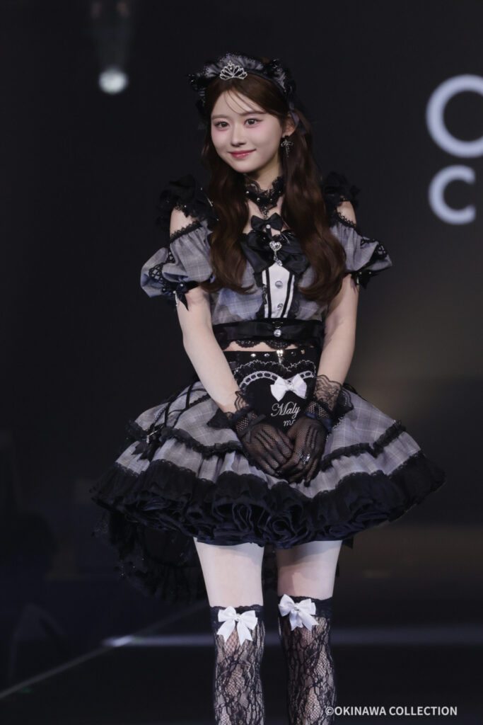 Young woman in a Gothic Lolita stage outfit with a tiara, lace gloves, and a plaid gray-and-black dress, posing on a dark stage.