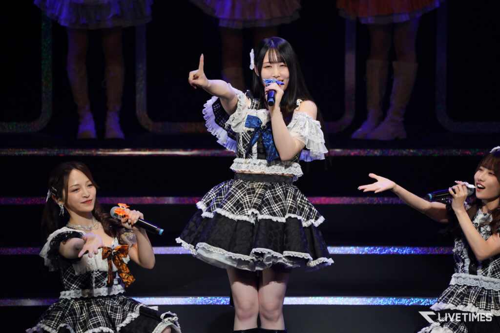 Center-stage idol singer points upward while holding a microphone during a group performance on a dark, glittery stage.