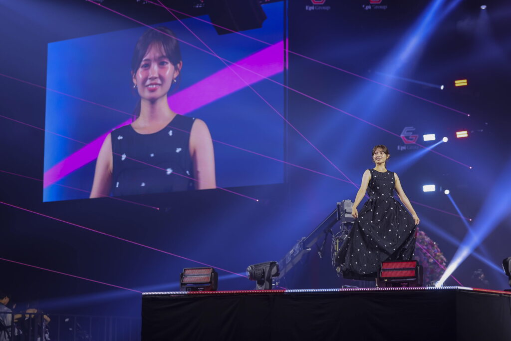 Stage performer walks down steps in a dark, polka-dot black dress as a large screen shows her portrait; pink laser lines and blue lighting fill the scene.