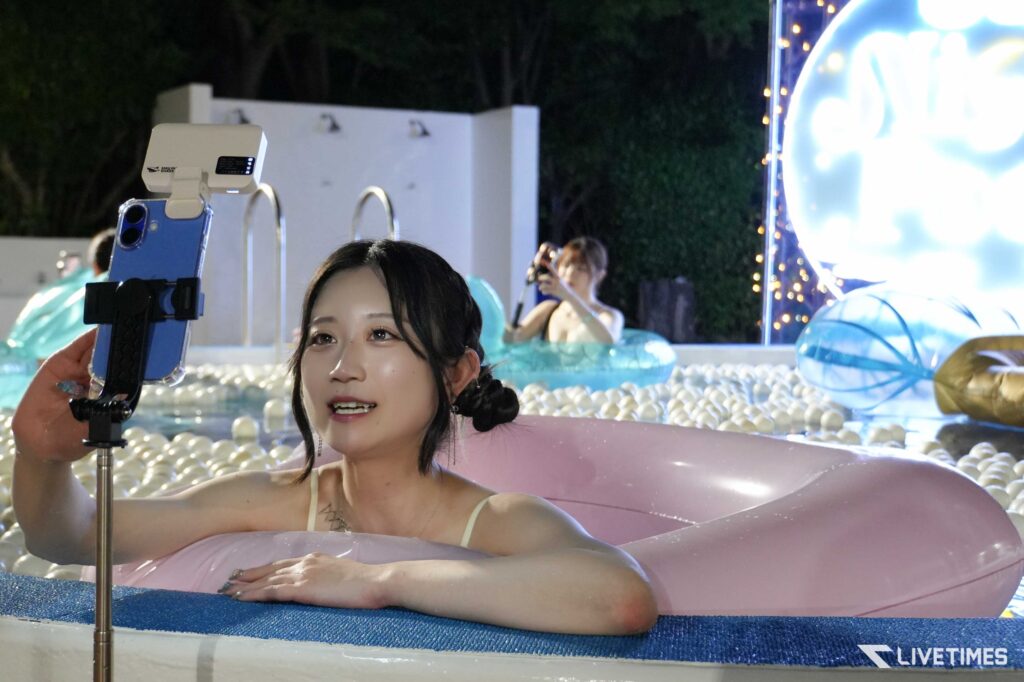 Woman in a pink inflatable pool sits at the edge, taking a selfie with a mounted smartphone on a selfie stick by the poolside.