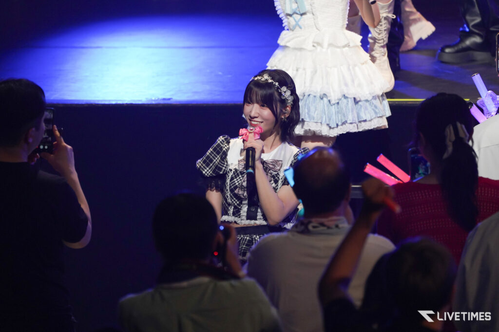 Female singer in a plaid costume with a pink bow microphone in hand, performing on a crowded stage.