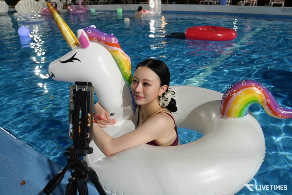 Woman in a burgundy swimsuit sits on a white unicorn inflatable float in a blue pool; in the foreground is a mounted action camera on a pole and a rainbow inflatable ring nearby.