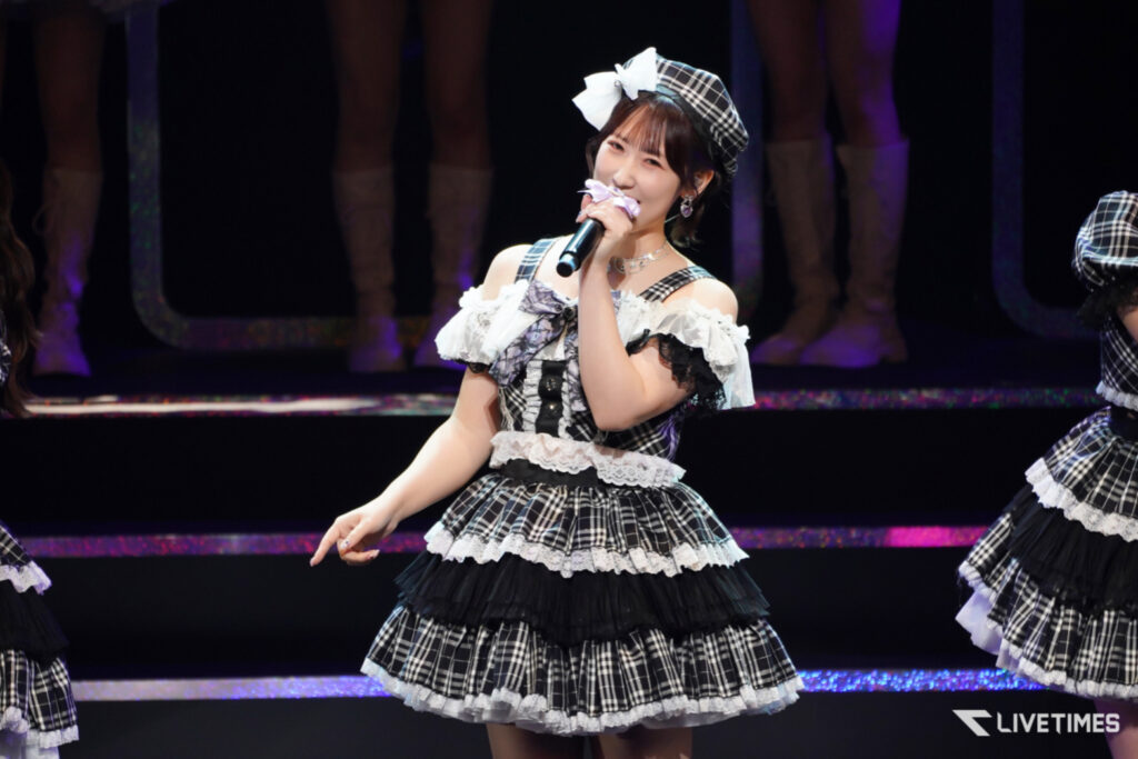 Woman in a black-and-white plaid frilly stage costume singing into a microphone with a bow headpiece during a performance.