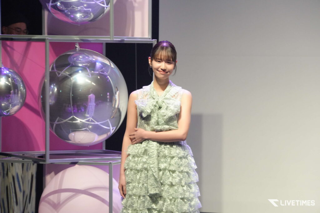 Young woman in a pale green ruffled dress standing on a stage beside large reflective spheres, smiling at the camera.