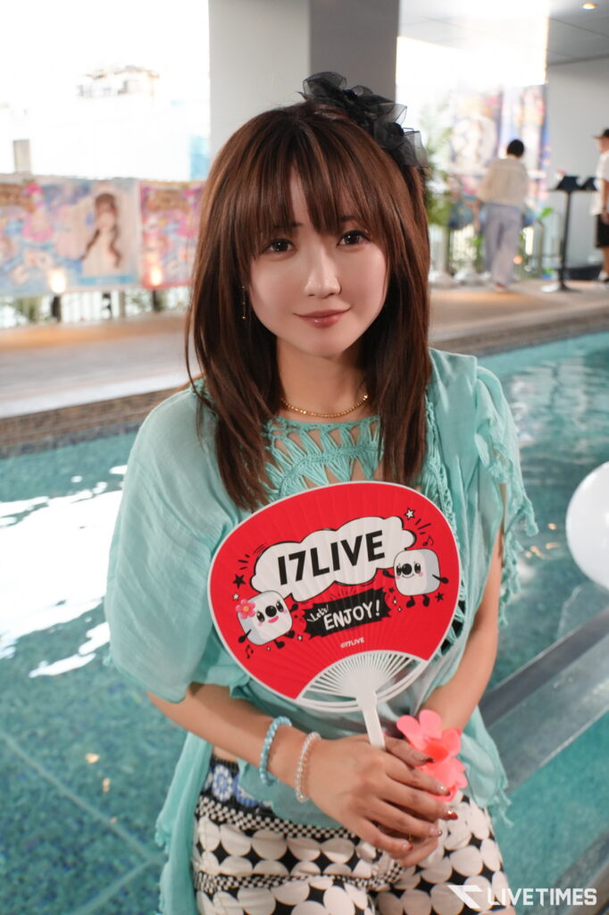 Smiling woman with brown hair and a black bow, wearing a turquoise top, holding a red IZLIVE fan by a poolside event venue