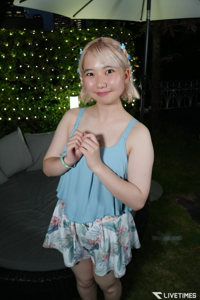 Smiling young girl with blonde hair in a blue sleeveless top, wearing blue flower clips, standing outdoors at night with fairy lights in the background and her hands clasped