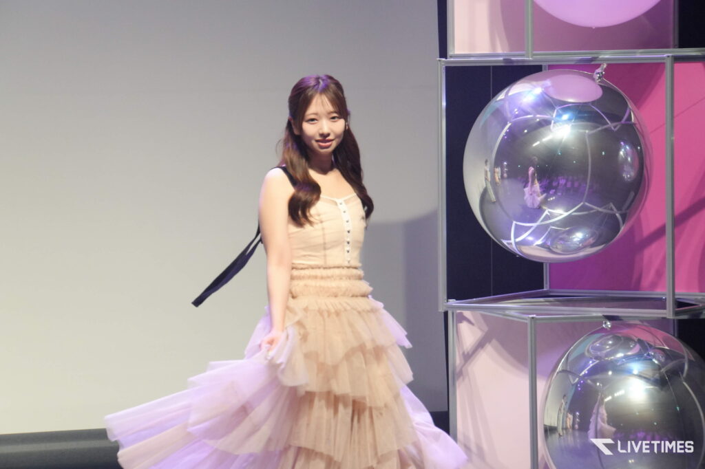 Young woman in a light beige sleeveless dress posing on a stage beside a display of large reflective glass spheres.",