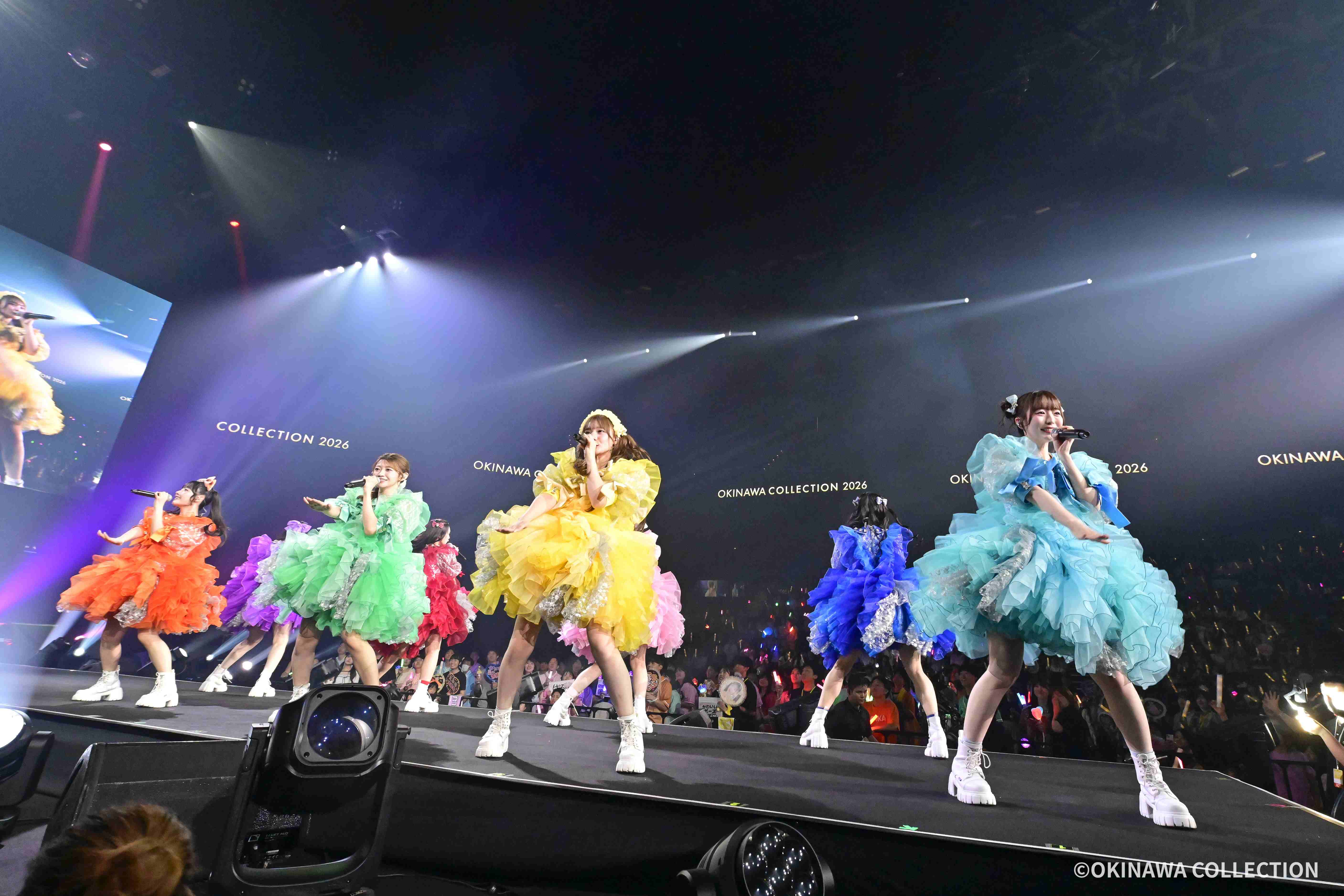 Group of female performers in bright, layered dresses performing on a stage labeled Okinawa Collection 2026.