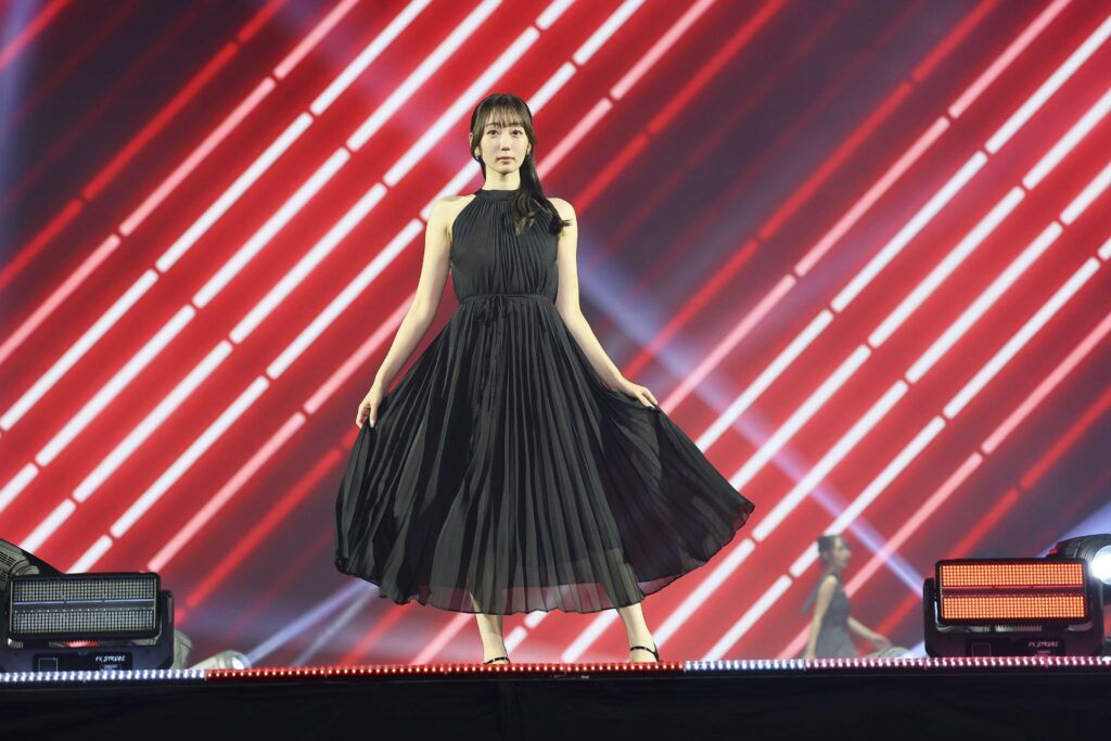 Woman in a flowing black gown performing center stage with a red diagonal-light backdrop behind her