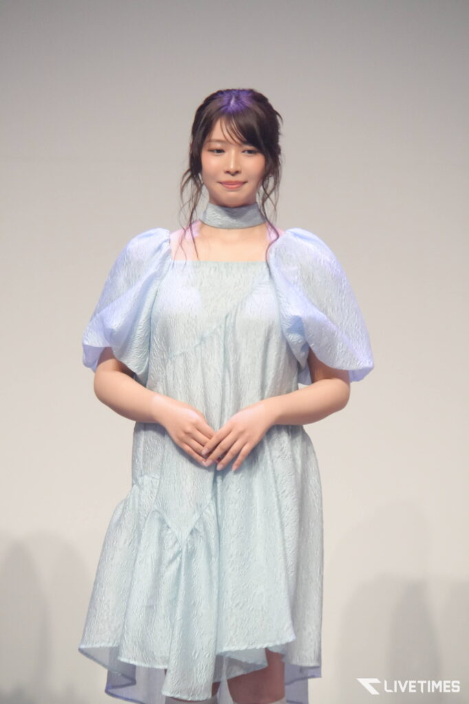 Young woman on stage wearing a pale blue, textured dress with puff sleeves and choker, hands gently clasped.