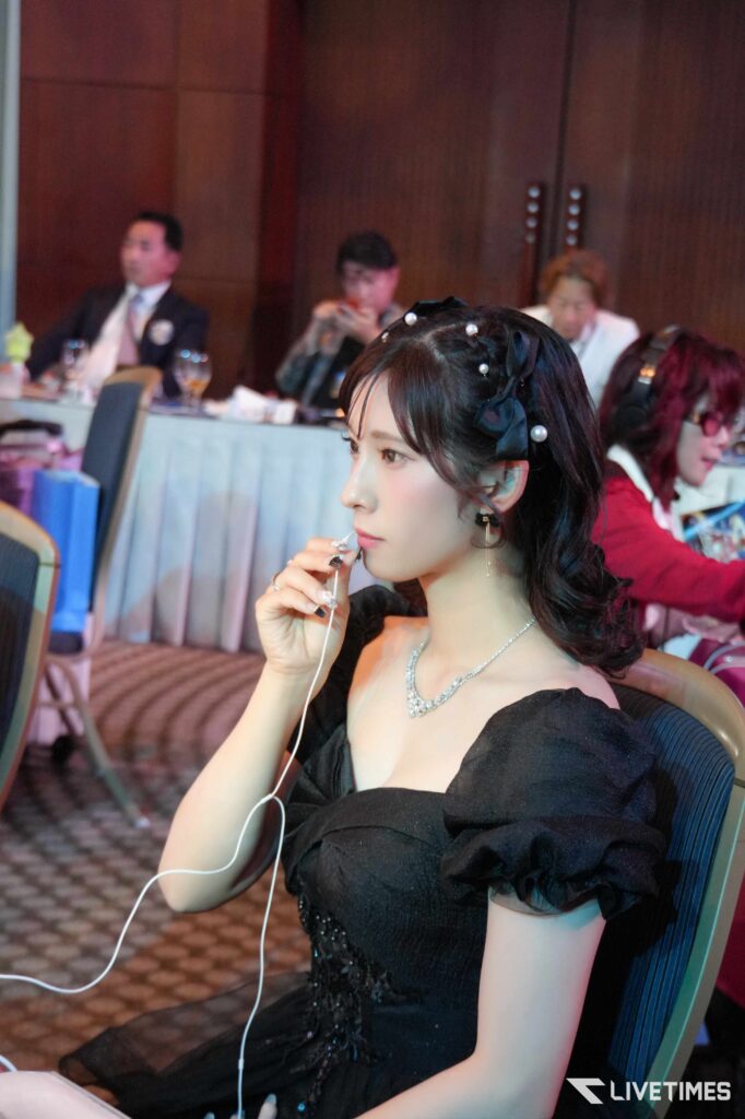 Young woman in a black formal dress with a pearl headband, drinking through a straw at a formal event, blurred background guests