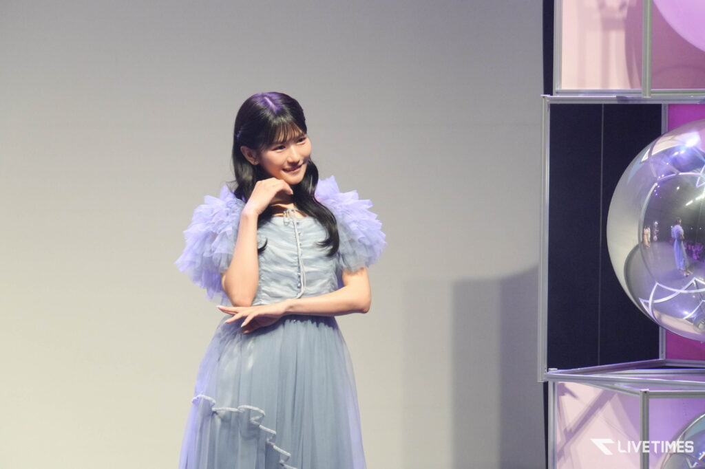 Young woman in a light blue, ruffled dress posing on a stage with a gradient backdrop and a glass display case lit with purple lighting on the right, smiling confidently.