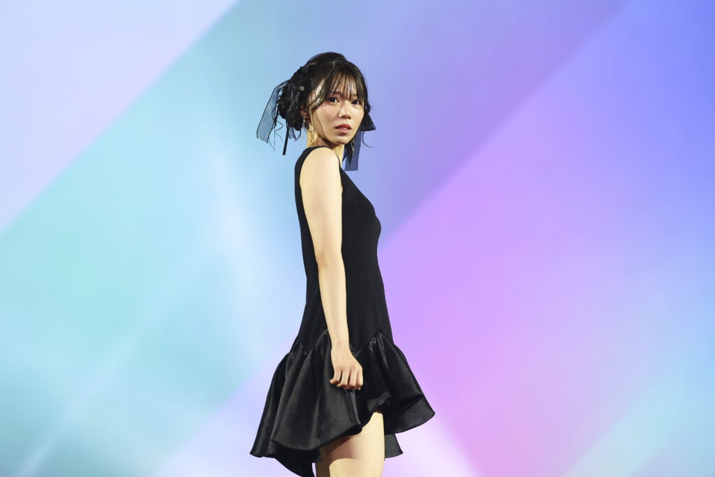 Woman in a black sleeveless dress posing sideways against a pastel gradient backdrop.