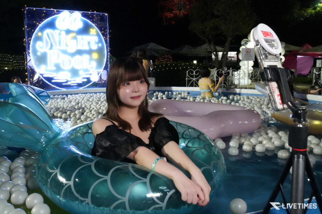 Young woman in a large teal inflatable float in a ball-pool at night, posing for a photo with neon lights in the background