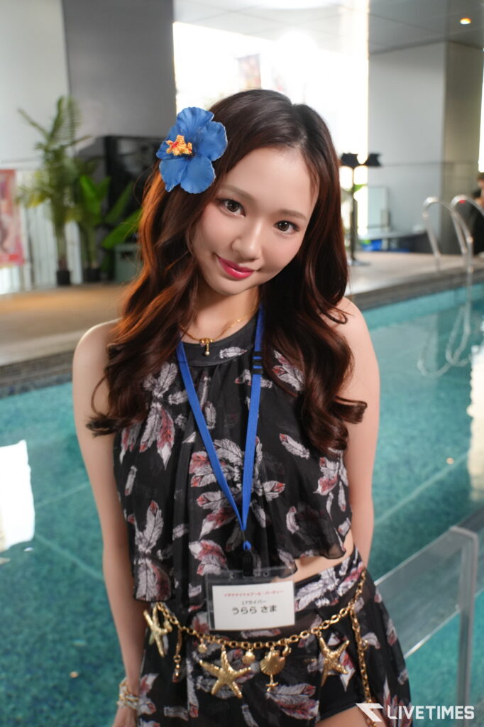 Young woman with a blue hibiscus in her hair, wearing a black floral dress and blue lanyard by an indoor pool area by glass railing.