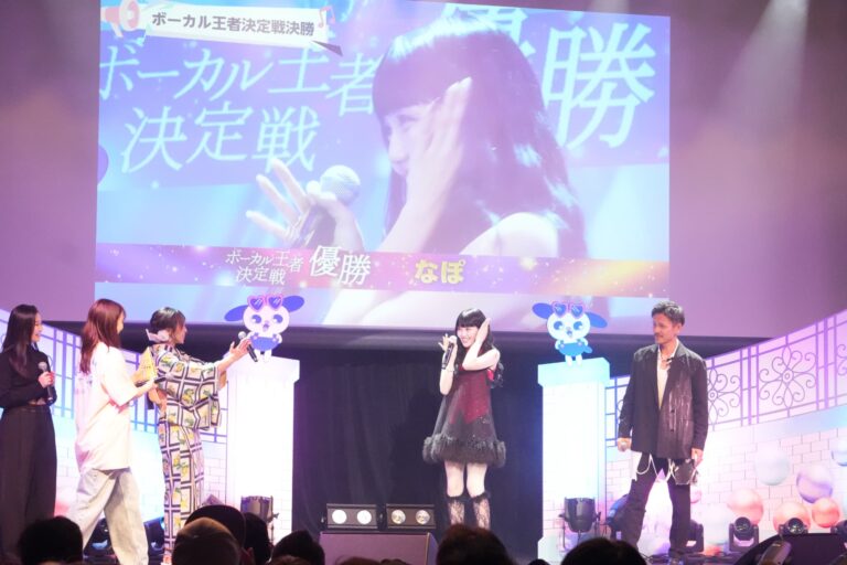 A female singer on stage with a microphone, flanked by hosts and a man, with a large screen displaying text behind them.