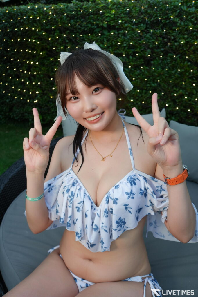 Smiling woman in a white and blue floral bikini, making peace signs with both hands, outdoors with string lights behind her.