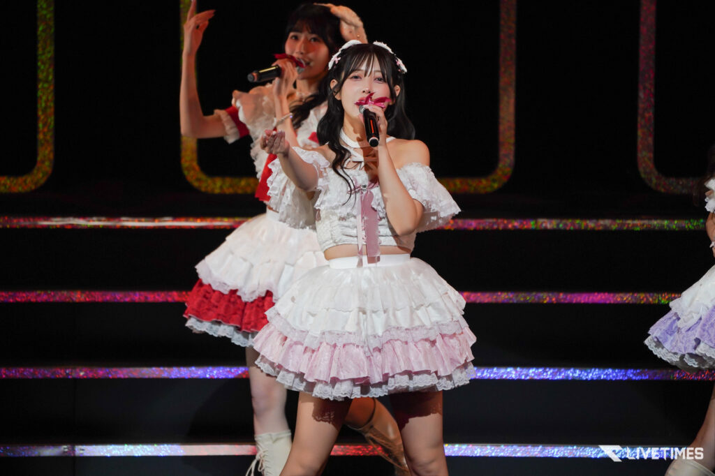 Two female singers in white frilly dresses perform on a dark stage with microphones and colorful stage lights behind them.