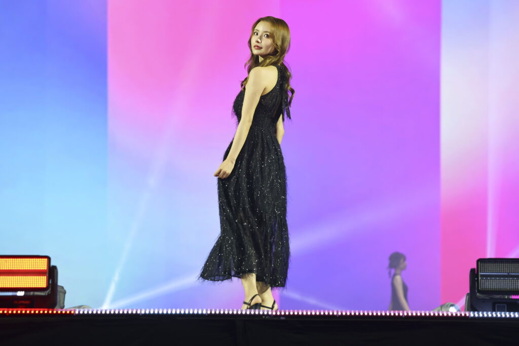 Female performer in a glittery black dress walking on stage against a blue-pink-purple gradient backdrop.