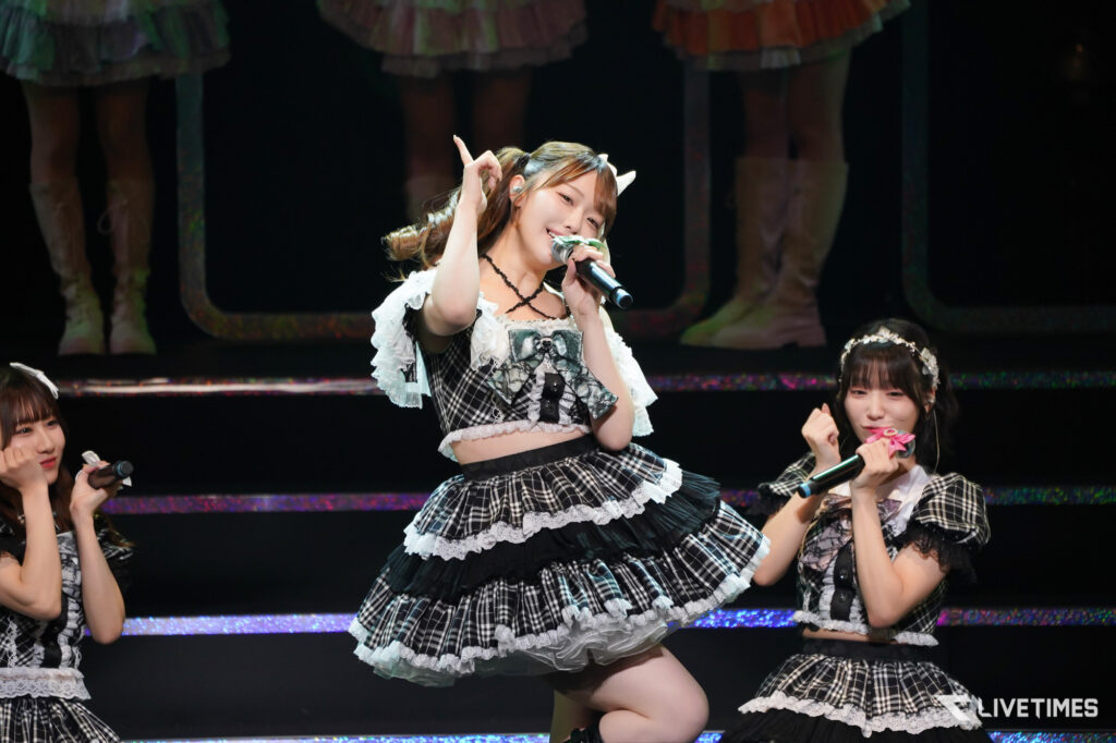 Group of young women in plaid stage outfits singing into microphones during a live performance on a dark stage with colorful lights behind them